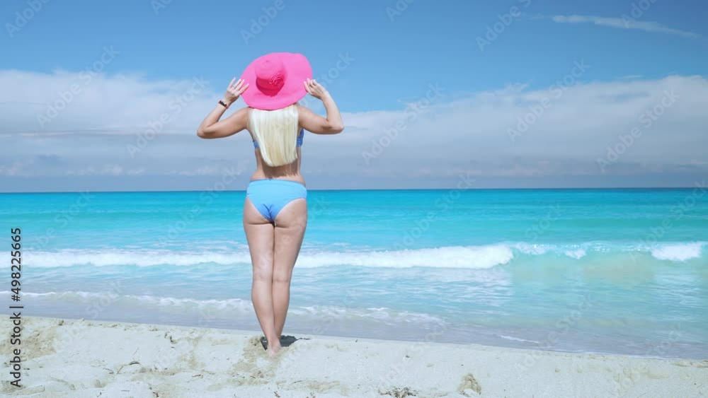 A young slender girl with blond long hair in a blue swimsuit stands on the ocean shore and holds a pink hat, enjoying the warm wind from the sea, 4K slow motion video. A young slender girl with blond long hair in a blue swimsuit stands on the ocean shore and holds a pink hat, enjoying the warm wind from the sea, 4K slow motion video.