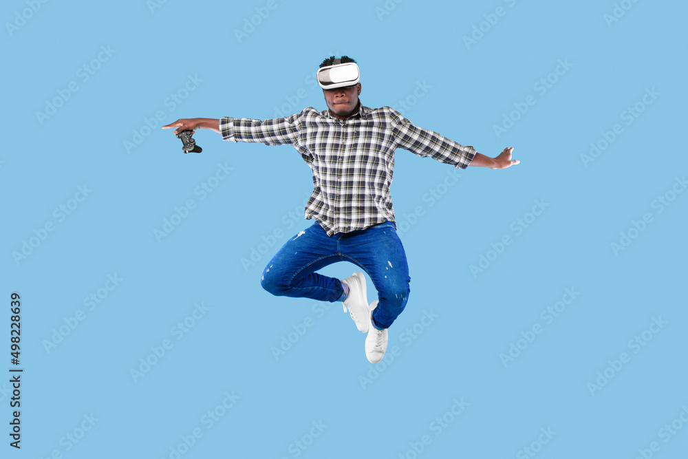 Young African American guy in VR headset holding joystick, jumping up in air on blue background