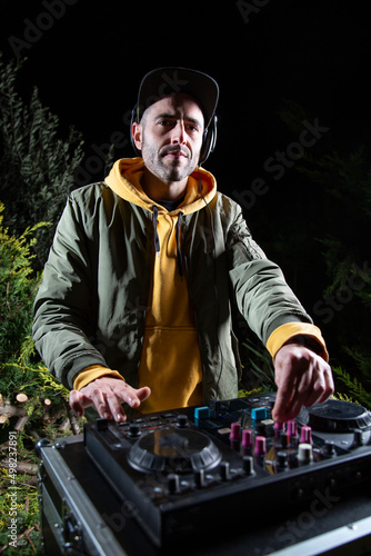 DJ vertical chico pincha musica con una mesa de mezclas en una fiesta en bosque vestido con una chaqueta verde, una sudadera con capucha y una gorra, rave party