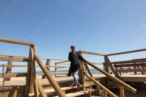A red-haired woman in a long black jacket, climbing some steps of a wooden walkway, on the beach of Isla Cristina, Spain.