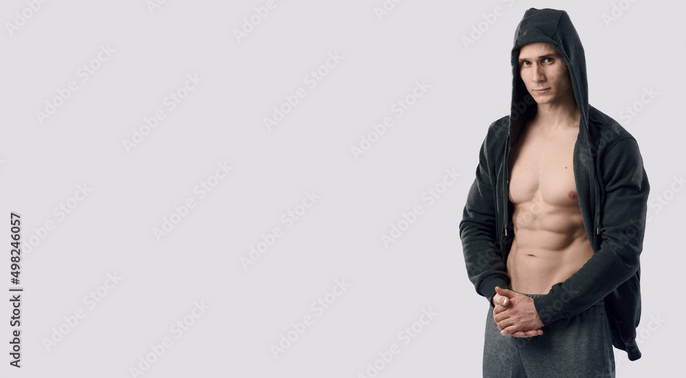 Caucasian muscular fitness model show body abs. Confident young fitness man with strong hands on gray background. Fitness man wearing hoodie on naked body. Caucasian muscular fitness model show body abs. Confident young fitness man with strong hands on gray background. Fitness man wearing hoodie on naked body.