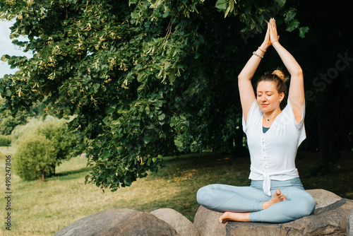 Meditation and yoga, mind and harmony concept. Smiling woman with closed eyes sitting in lotus position with arms raised up in nature, copy space. Yogi woman meditating outdoors
