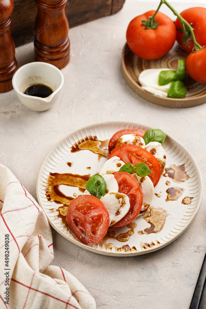 Traditional italian salad caprese consisting of slices red tomatoes, fresh basil and mozzarella cheese with balsamic vinegar dressing on concrete background, wooden pepper and salt mills Traditional italian salad caprese consisting of slices red tomatoes, fresh basil and mozzarella cheese with balsamic vinegar dressing on concrete background, wooden pepper and salt mills