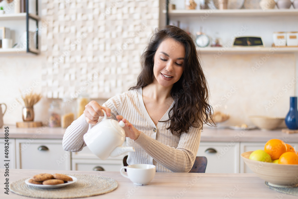 Cheerful woman in casual pouring tea in cup, sitting at table in kitchen interior, enjoying morning