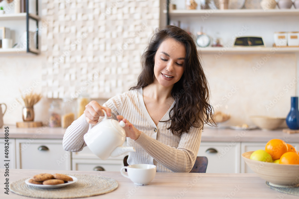 Cheerful woman in casual pouring tea in cup, sitting at table in kitchen interior, enjoying morning Cheerful woman in casual pouring tea in cup, sitting at table in kitchen interior, enjoying morning
