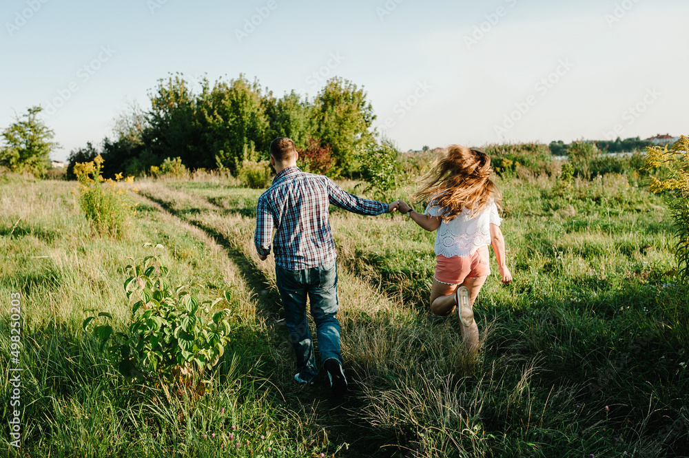 Fototapeta premium Rear view of a romantic man and woman walking on field grass, nature enjoying sunlight. Concept of lovely family holding hands. Young couple running and looking away. Back view.