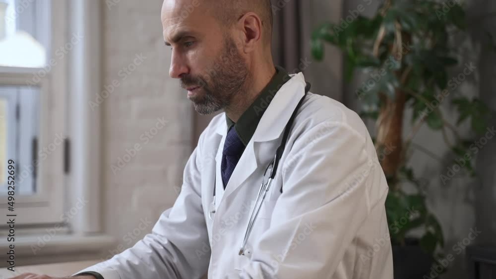 Portrait of male doctor typing diagnosis on laptop and sitting at table in modern clinic spbas. Close view of middle-aged man looks at computer screen and types on keyboard, does diagnostic work and Portrait of male doctor typing diagnosis on laptop and sitting at table in modern clinic spbas. Close view of middle-aged man looks at computer screen and types on keyboard, does diagnostic work and