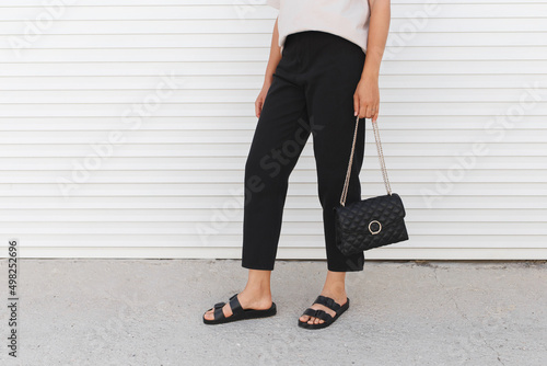 Woman wearing beige t-shirt, black pants, bag and flat sandals walking outdoor near white roller door. Details of stylish trendy basic minimalistic casual outfit. Street fashion. Women's legs, no face