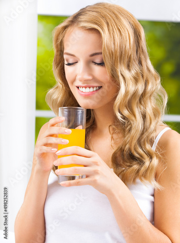 Portrait image of young happy smiling woman with closed eyes, hjlding glass of orange juice near window, at home. Amazed blond girl indoors. Healthy eating and dieting concept.