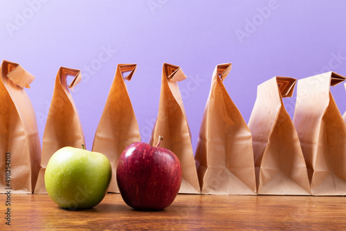 Tela Apple and granny smith apple by row of paper lunch bags on table against purple