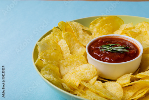 Close-up of red sauce in bowl amidst potato chips served in plate on blue background