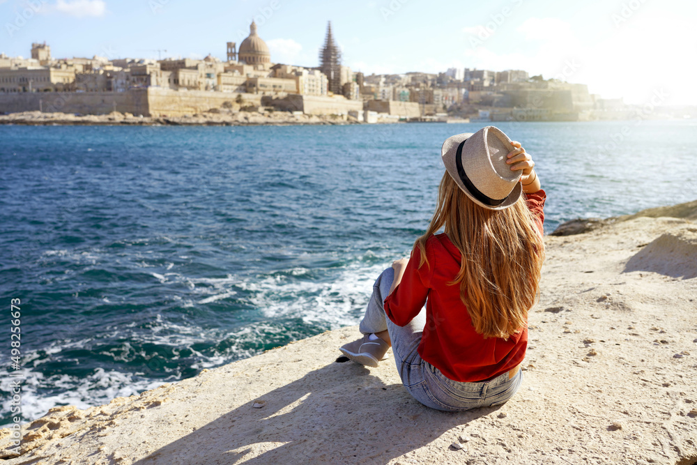 Obraz na płótnie Beautiful young woman with hat sitting on stone by the sea looking at stunning panoramic view of Valletta city in Malta w salonie
