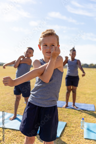 Multiracial elementary schoolboys doing stretching exercise while standing on yoga mat against sky