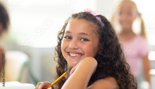 Portrait of smiling biracial elementary schoolgirl sitting with hand on chin in classroom
