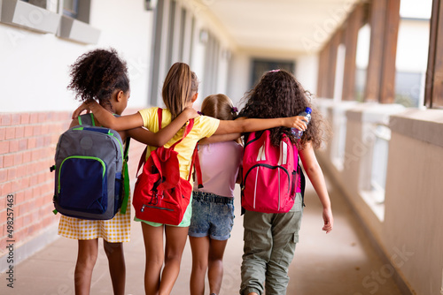 Rear view of multiracial elementary schoolgirls with backpacks and arm around walking in corridor photo
