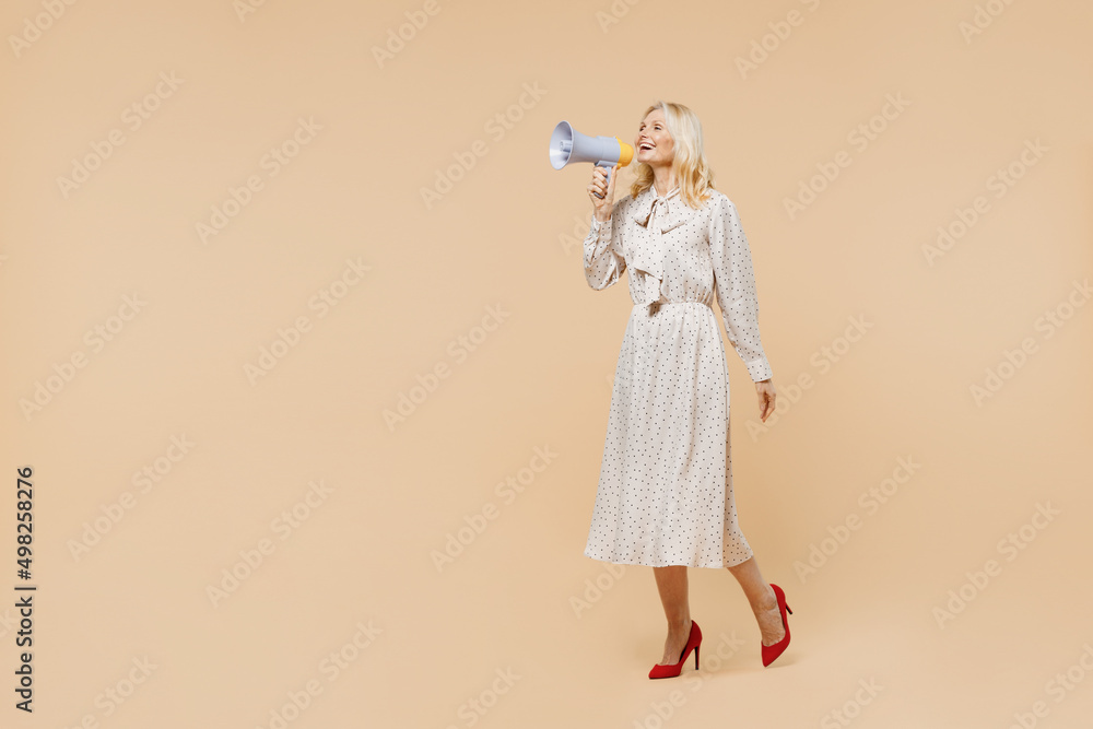Full size body length promoter excited elderly gray-haired blonde woman lady 40s years old wears pink dress go move hold scream in megaphone isolated on plain pastel beige background studio portrait. Full size body length promoter excited elderly gray-haired blonde woman lady 40s years old wears pink dress go move hold scream in megaphone isolated on plain pastel beige background studio portrait.