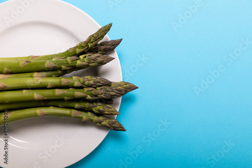 Overhead view of raw asparagus in white plate by copy space over blue background