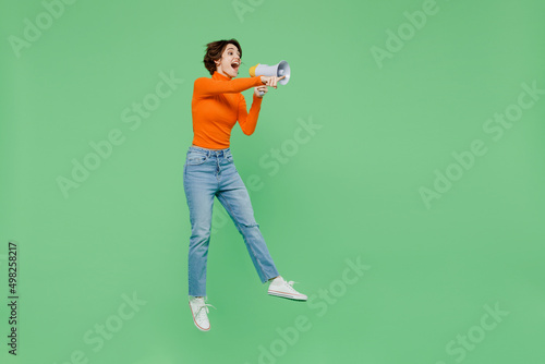 Full body young woman 20s wear orange turtleneck hold scream in megaphone announces discounts sale Hurry up jump high point finger aside on workspace isolated on plain pastel light green background.