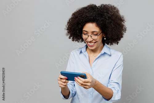 Young fun employee business corporate lawyer woman of African American ethnicity in classic shirt work in office use play racing app on mobile cell phone gadget smartphone isolated on grey background.