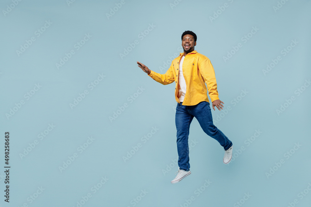 Full body side view young man of African American ethnicity 20s wear yellow shirt jump high walking going run isolated on plain pastel light blue background studio portrait. People lifestyle concept.
