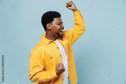 Fotografiet Side view young fun man of African American ethnicity in yellow shirt doing winner gesture celebrate clenching fists say yes isolated on plain pastel light blue background