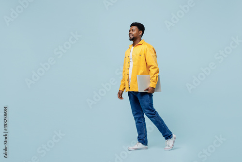Full body young man of African American ethnicity 20s wearing yellow shirt hold use work on laptop pc computer isolated on plain pastel light blue background studio portrait. People lifestyle concept.