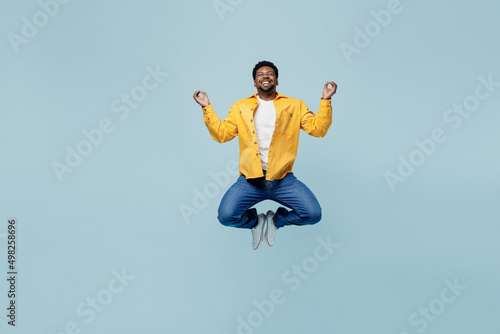 Full body young man of African American ethnicity in yellow shirt jump high hold spreading hands in yoga om aum gesture relax meditate try to calm down isolated on plain pastel light blue background