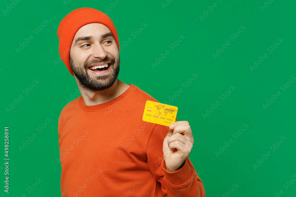 Young smiling cheerful cool happy caucasian man 20s wear orange sweatshirt hat hold in hand credit bank card look aside on workspace isolated on plain green background studio People lifestyle concept