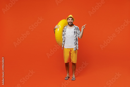 Full body young tourist man in beach shirt hat hold inflatable ring point index finger aside on workspace isolated on plain orange background studio portrait Summer vacation sea rest sun tan concept