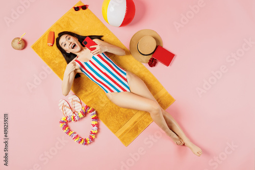 Top view full size woman of Asian ethnicity in striped swimsuit lies on towel hotel pool use point finger mobile cell phone isolated on plain pink background. Summer vacation sea rest sun tan concept.
