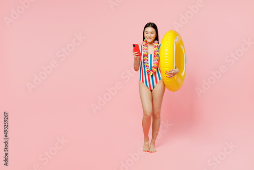 Full body young woman of Asian ethnicity in striped one-piece swimsuit lei hold inflatable ring mobile cell phone isolated on plain pastel pink background. Summer vacation sea rest sun tan concept