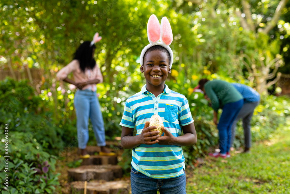 Naklejka premium Portrait of happy african american boy in bunny ears holding easter egg while family in backyard