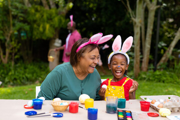 Cheerful african american girl and grandmother wearing bunny ears while painting eggs on easter day