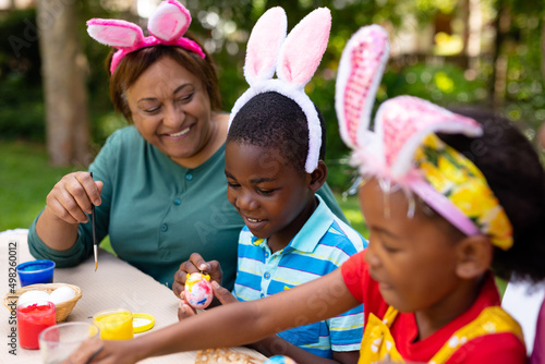 Happy african american siblings and grandpmother in bunny ears painting easter eggs in backyard