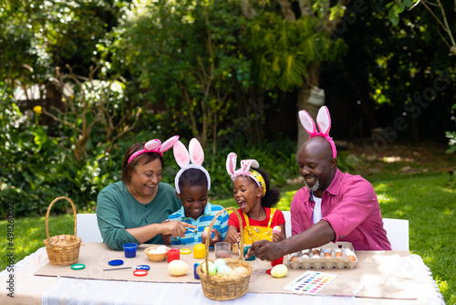 Cheerful african american siblings and grandparents in bunny ears painting eggs on easter day