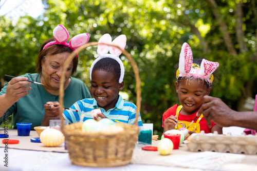 Smiling african american siblings and grandmother wearing bunny ears while painting eggs on easter