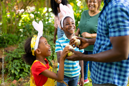 Happy african american siblings in bunny ears giving easter eggs to father in backyard
