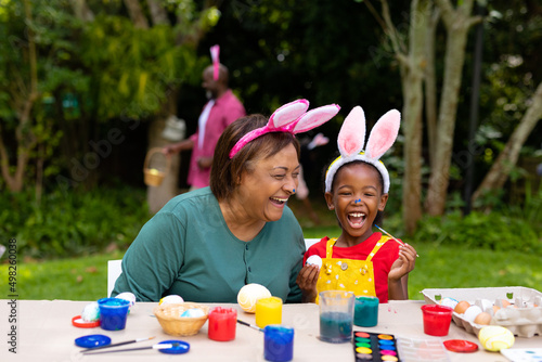 Cheerful african american girl and grandmother wearing bunny ears while painting eggs on easter day