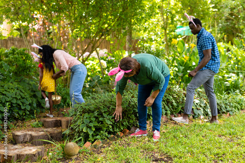 African american multi-generational family in bunny ears searching easter eggs in backyard