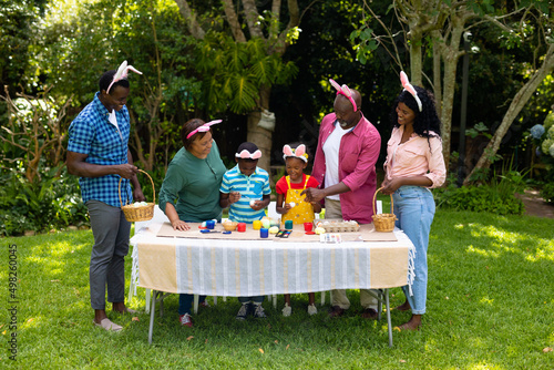 Happy african american multigenerational family in bunny ears painting easter eggs in backyard