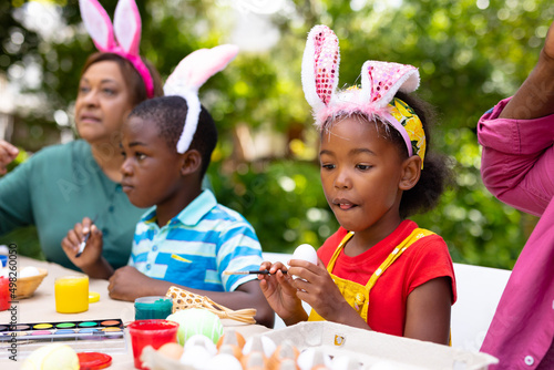 African american girl wearing bunny ears painting eggs with family in backyard on easter day