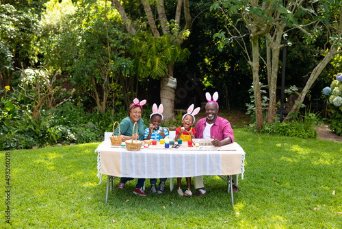 Happy african american siblings and grandparents in bunny ears painting easter eggs in backyard