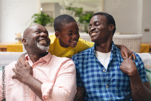 Happy african american boy with father and grandfather sitting on sofa at home