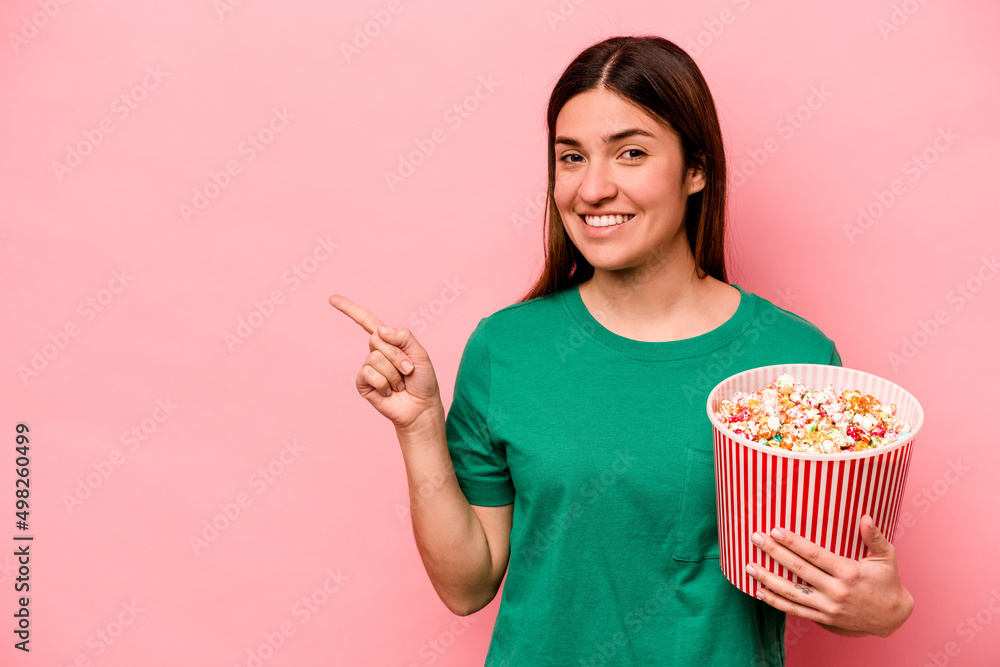 Young caucasian woman holding popcorn isolated on pink background smiling and pointing aside, showing something at blank space.