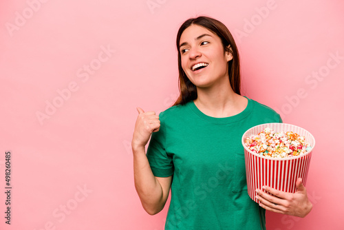 Young caucasian woman holding popcorn isolated on pink background points with thumb finger away, laughing and carefree.