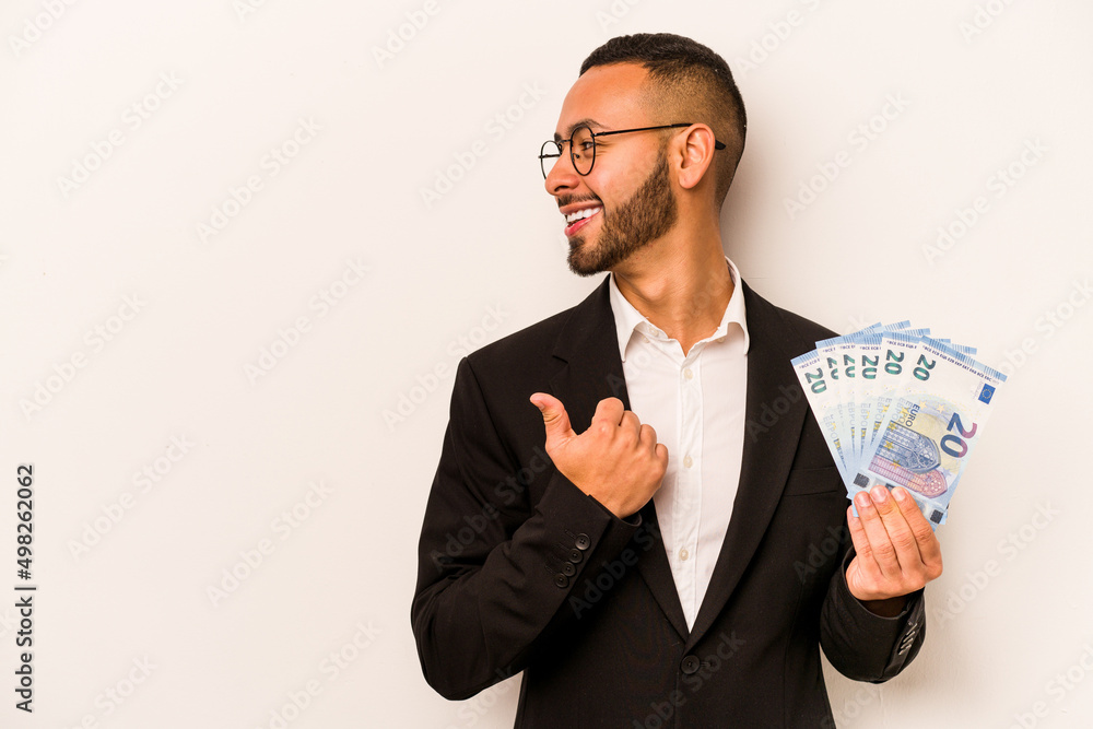 Young business hispanic man holding banknotes isolated on white background points with thumb finger away, laughing and carefree. Young business hispanic man holding banknotes isolated on white background points with thumb finger away, laughing and carefree.