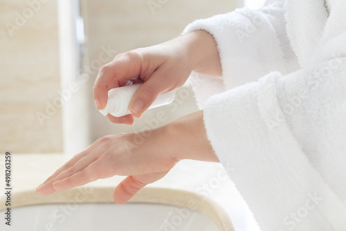 .Woman in the robe applying cream after washing hands for protective and care dry skin near white sink in bathroom with sunlight from the window.