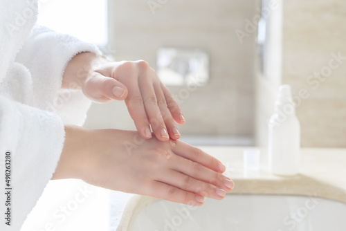 Woman in the robe applying cream, gel, serum after washing hands for protective and care dry skin near white sink in bathroom with sunlight from the window.