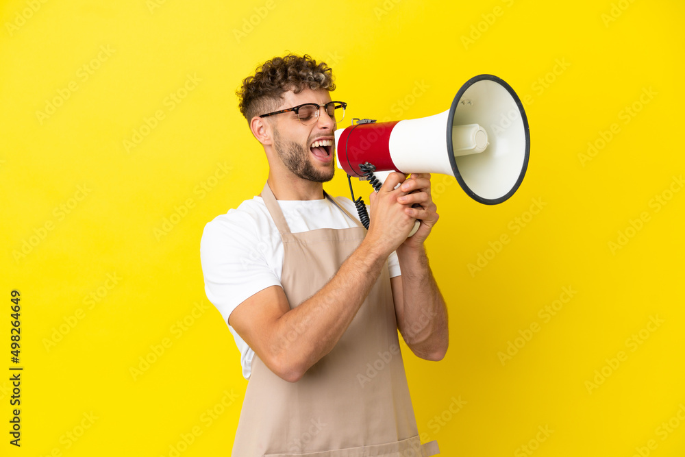 Restaurant waiter blonde man isolated on yellow background shouting through a megaphone Restaurant waiter blonde man isolated on yellow background shouting through a megaphone