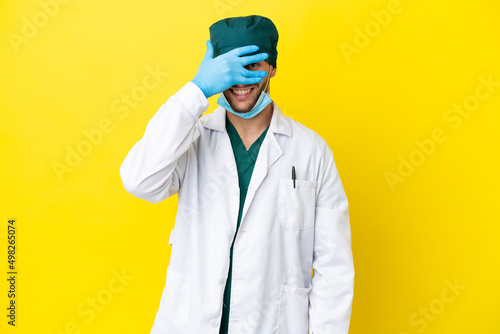 Surgeon blonde man in green uniform isolated on yellow background covering eyes by hands and smiling